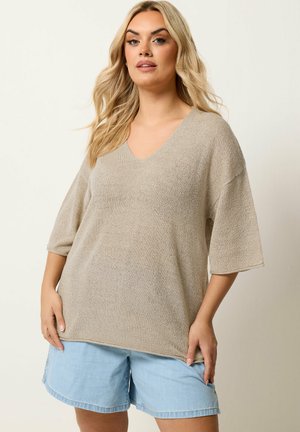 Blonde Frau trägt einen beigen, strukturierten, locker sitzenden V-Ausschnitt-Pullover mit ellbogenlangen Ärmeln und hellblauen Jeansshorts.