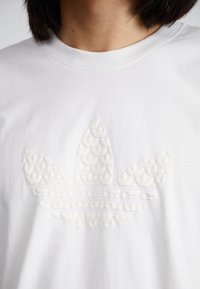 Balta medvilnė marškinėliai su tekstūruota, tonine Adidas logotipu ant krūtinės, su trimis lapo formos elementais ir lygiagrečiais raštais žemiau.