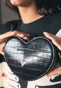 Melnas sirds formas soma no lakādas ar krokodilāda reljefu. Aprīkota ar zelta detaļām un reljefuzogo "ROCKETT GEORGE."