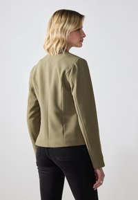 Olivgrön skräddarsydd jacka med långa ärmar, strukturerade axelsömmar och en figursydd design. Slät tyg, inga synliga knäppningar eller mönster.