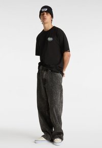 Black short-sleeve t-shirt with a graphic logo, paired with loose-fitting gray jeans and beige sneakers. Accessorized with a black beanie.