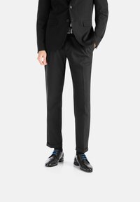 Schwarze Anzughose mit taillierter Passform, kombiniert mit glänzenden schwarzen Dressschuhen mit blauen Schnürsenkeln und schwarzen Socken. Glatte Stofftextur sichtbar.