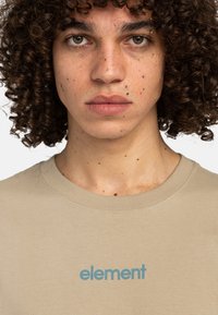 Beige bomulds t-shirt med rund halsudskæring og et blåt "element" logo trykt på brystet. Tekstureret stof med naturlige sprøjt.