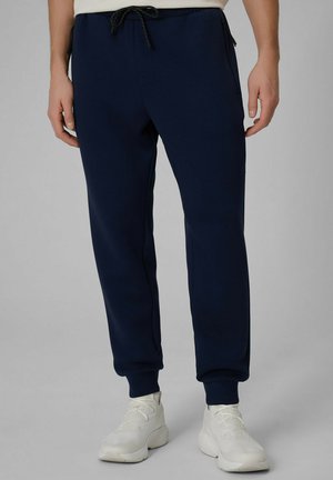 Mężczyzna ubrany w granatowe spodnie dresowe ze ściągaczem w talii i białe trampki, stojący na jednolitym tle.