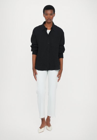 Camisa negra texturizada de botones con un bolsillo frontal, combinada con jeans cortos de color azul claro y mules beige, mostrando una paleta de colores neutros.