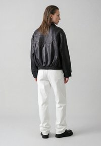 Person med vådt, glat tilbagekammet hår iført en sort læderjakke med "VERTERE BERLIN" på bagsiden, hvide bukser og sorte sko, står i profil.