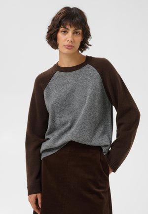 Kvinde med kort mørkt hår iført grå og brun langærmet sweater og brun nederdel, stående mod en ensfarvet lys baggrund.