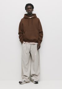 Brown hooded sweatshirt with front pocket, layered over a white T-shirt with red accents, paired with wide grey trousers and patterned sneakers.