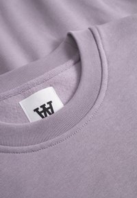 Light purple crewneck sweatshirt with ribbed collar and cuffs, featuring a fabric label with black text. Soft, textured finish.