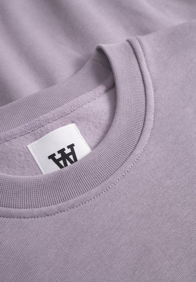 Sweatshirt violet clair à col rond avec un col et des poignets en côtes, présentant une étiquette en tissu avec du texte noir. Fini doux et texturé.