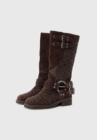 Stivali al ginocchio in suede marrone con borchie decorative, cinghie a doppia fibbia e punta rotonda su un tacco basso impilato.