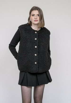 Femme aux cheveux mi-longs portant un gilet noir texturé boutonné, un haut à manches longues et une jupe plissée, debout devant un fond uni.