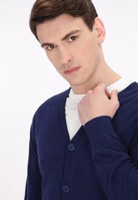Mornarsko modra cardigan iz mehkega blaga, s polo ovratnikom, dvema modrima gumboma in rebrastimi manšetami, oblečen preko bele srajce.