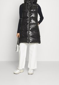 Czarna pikowana kamizelka o połyskującym wykończeniu, z zamkiem i kapturem. Noszona na dopasowanej czarnej bluzce z długim rękawem oraz białych szerokich spodniach.