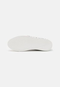 Semelle de sneaker blanche avec un motif de grille texturé et une zone de talon lisse, photographiée par le dessous sur un fond uni.