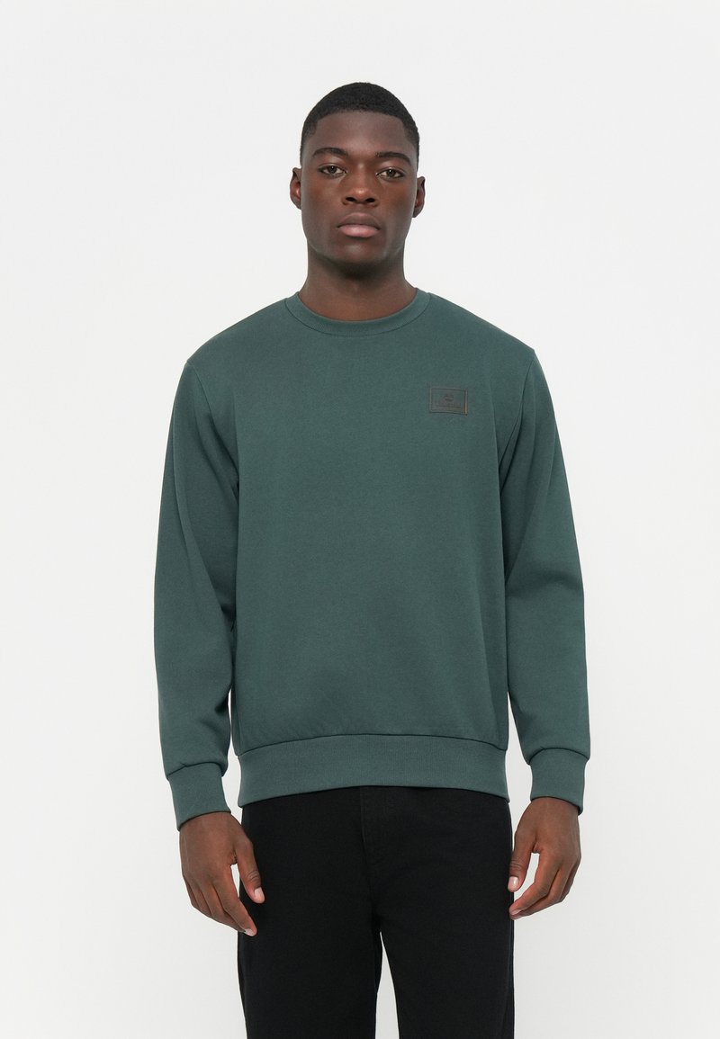 Donkergroen sweatshirt met een ronde hals en lange mouwen. Beschikt over een rechthoekige stoffen patch op de borst. Gladde textuur, ontspannen pasvorm.