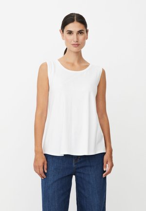 Femme aux cheveux foncés attachés, portant un haut blanc sans manches et un jean bleu, debout devant un fond blanc uni.