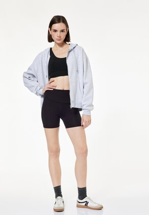 Junge Frau trägt schwarzen Sport-BH, schwarze Radlerhose, grauen Kapuzenpullover mit Reißverschluss, dunkle Socken und weiße Sneakers, steht mit der Hand in der Hüfte.