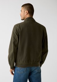 Lekka oliwkowo-zielona kurtka z prostym kołnierzem, ściągaczami przy rękawach i w pasie. Posiada tylne szwy i luźny krój.