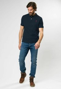Marineblå polo t-shirt med krave, korte ærmer og et rødt logo. Sættes sammen med blå jeans og brune snørebåndsstøvler. Glat stofstruktur.