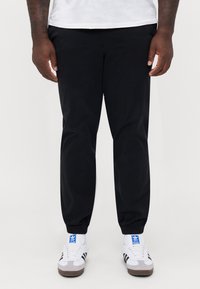 Sorte jogger-bukser med elastiske manchetter, lavet af et glat stof. Kombineret med hvide og grå sneakers med sorte striber.
