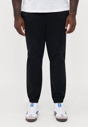 Zwarte joggings met elastische boorden, gemaakt van een gladde stof. Gepaard met wit en grijze sneakers met zwarte strepen.