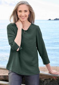 Lächelnde Frau mit grauem Haar in einer dunkelgrünen Langarmbluse posiert an einer Steinmauer und einem Metallgeländer mit Ozean und Himmel im Hintergrund.