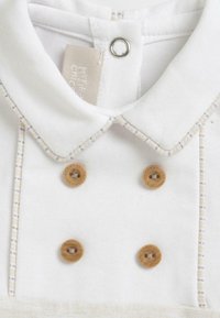 Chemise blanche à col avec des boutons en bois, en tissu doux, et des accents beiges sur le col. Présente une étiquette près de l'encolure.