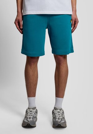 Man die turquoise knielange shorts draagt, witte sokken, grijze en witte sneakers en een wit shirt, staand tegen een effen achtergrond.