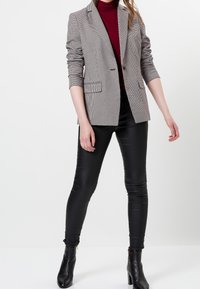 Blazer à carreaux noir et blanc avec un revers cranté, deux poches avant, superposé à un col roulé bordeaux, associé à des leggings noirs et des bottines.