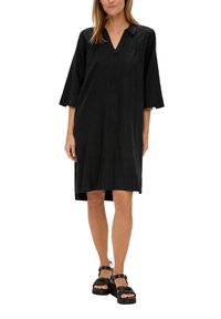 Zwart linnen jurk met een V-hals, driekwartmouwen en zijsplitten. Complementen een model die zwarte platform sandalen draagt.