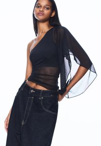 Zwarte een-schouder top met doorschijnende overlaagmouw, gecombineerd met donkerblauwe hooggetailleerde spijkerbroek met contrasterende naden en zakken.