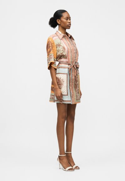 Γυναίκα που στέκεται φορώντας πολύχρωμο, με σχέδια φόρεμα με ζώνη, κοντά μανίκια και μπεζ πέδιλα με ψηλό τακούνι, μπροστά σε απλό φόντο.