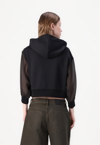 Μαύρο κοντό φούτερ με κουκούλα, που διαθέτει διάφανες μαύρες φουσκωτές μανσέτες. Συνδυάζεται με ελιόχρωμες παντελόνες ψηλής μέσης με πίσω τσέπες.