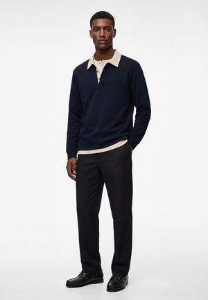 Młody mężczyzna stoi, ubrany w granatową koszulkę polo z długim rękawem na beżowej koszuli, czarne spodnie i czarne eleganckie buty, na tle jednolitej białej ściany.