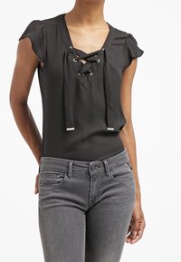 Schwarze Bluse mit V-Ausschnitt und Schnürung vorn. Kurzärmel mit Rüschen. Gepaart mit eng anliegenden grauen Jeans, die eine subtile Textur hinzufügen.