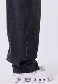 Jeans in denim neri con vestibilità dritta, dotati di cucitura laterale e orlo leggermente strappato, abbinati a scarpe da ginnastica bianche.