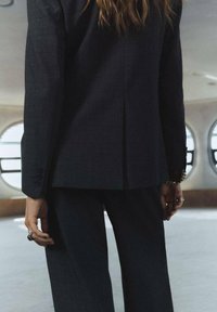 Chaqueta y pantalones de traje de cuadros oscuros, diseño a medida, abertura trasera visible, botones negros minimalistas, mano del modelo adornada con anillos.