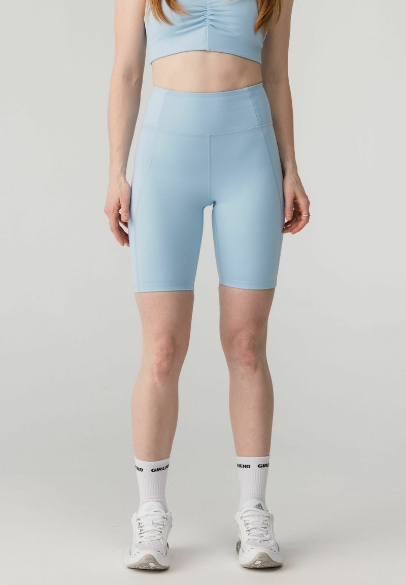 Femme portant un short cycliste taille haute bleu clair, un débardeur assorti raccourci, des chaussettes blanches avec un texte, et des chaussures de sport blanches, vue de face.
