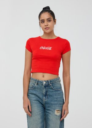 T-shirt rossa corta realizzata in cotone, con maniche corte e scollo rotondo, presentante il logo bianco della Coca-Cola sul davanti. Indossata con jeans in denim strappati.