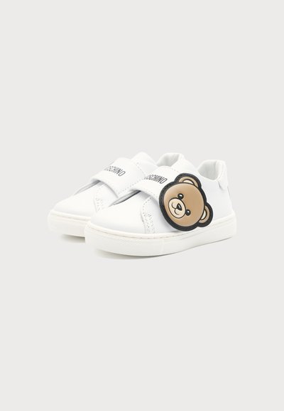 Baskets blanches pour enfants avec des bandes Velcro et un visage d'ours en peluche marron sur le côté d'une chaussure.
