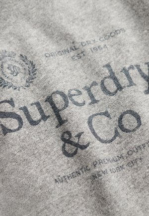 Tessuto grigio con grandi scritte nere "Superdry & Co." e ulteriori piccoli caratteri tipografici e un dettaglio di logo circolare.