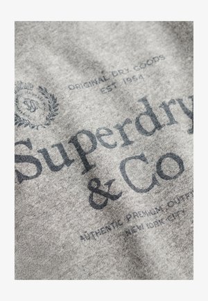 Tessuto grigio con grandi scritte nere "Superdry & Co." e ulteriori piccoli caratteri tipografici e un dettaglio di logo circolare.