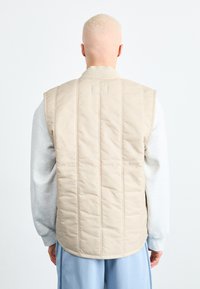 Vest bej cu umplutură, cu guler texturat și fără mâneci, văzut din spate, asortat cu o cămașă cu mânecă lungă de culoare gri deschis și pantaloni blue.