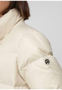 Žena v krémovej pufrovacej bunde s vysokým golierom a malým čiernym kruhovým logom na hornom ramene na šedom pozadí.