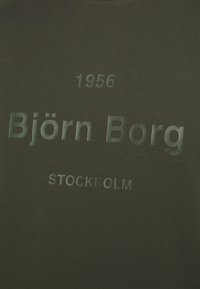 Olivově zelené tričko s hladkou texturou. Obsahuje vystouplý text: "Björn Borg", "1956" a "STOCKHOLM" v kontrastním písmu.
