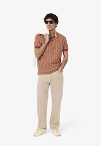 Brunmönstrad polo skjorta med röd krage, beige byxor och gula sneakers; bär solglasögon med en vit tröja hängande över axeln.