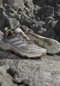 Chaussures de randonnée beige et blanches avec semelles robustes et marquage Gore-Tex sur terrain rocheux.