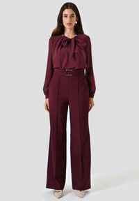 Blouse bordeaux avec un grand nœud au cou et des manches longues, associée à un pantalon large taille haute et des chaussures de couleur claire.