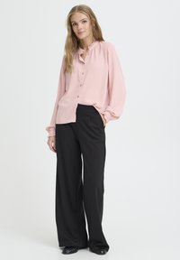Blouse rose pâle avec un col à volants et une fermeture à boutons, associée à un pantalon large taille haute noir et des chaussures noires.